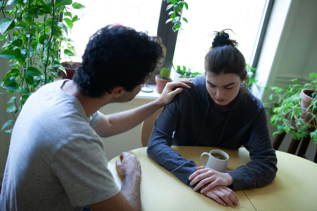 A teen looking down at a mug of tea sits with a concerned adult at a table.