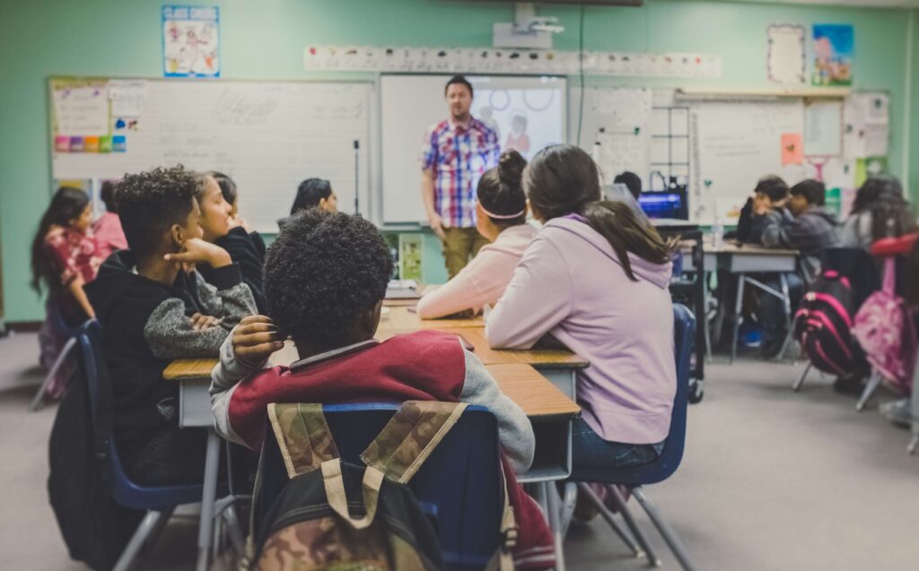 A group of students sit in a brightly-lit classroom looking toward their teacher at the front of the room.