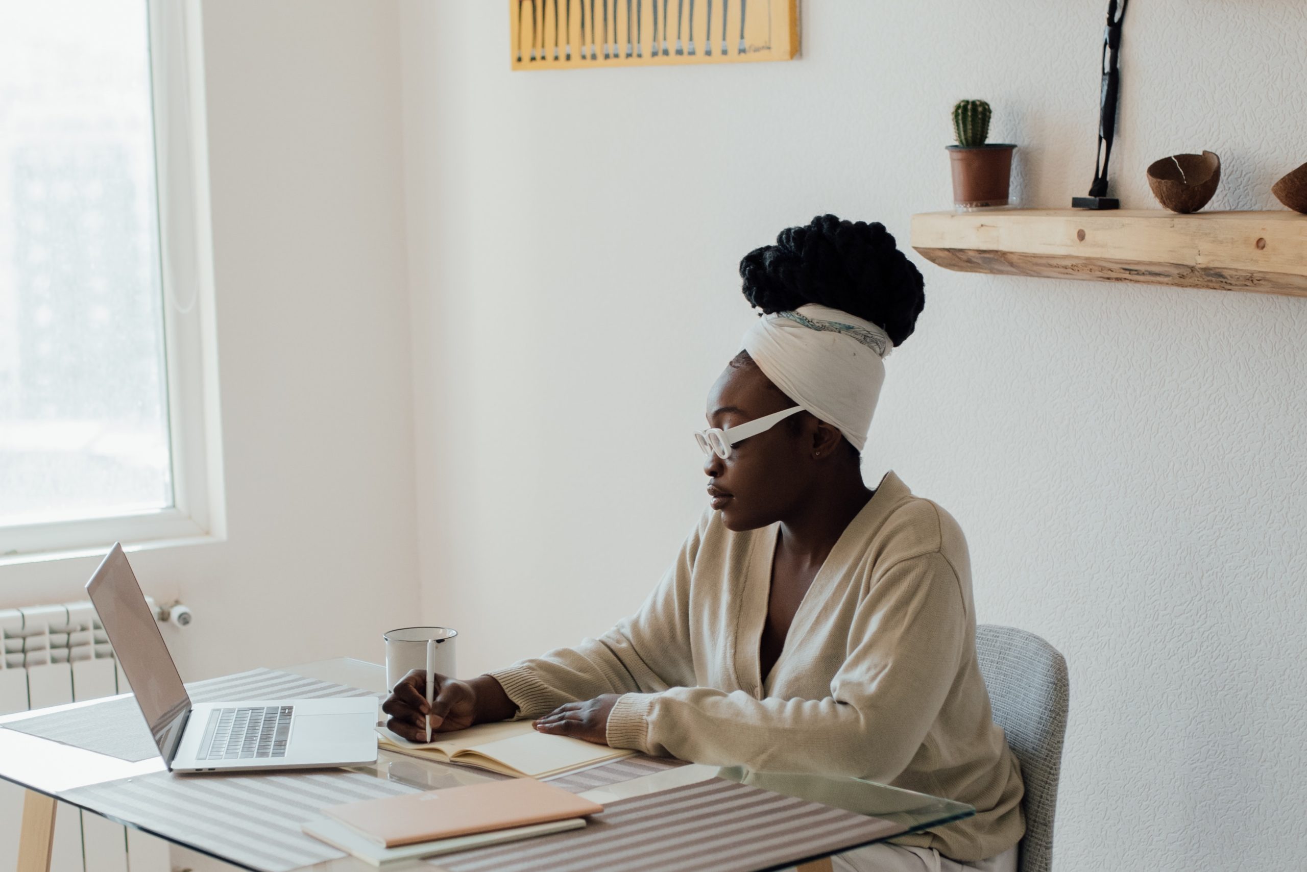 woman wearing a headwrap sitting at a desk writing in a journal with a laptop open in front of her