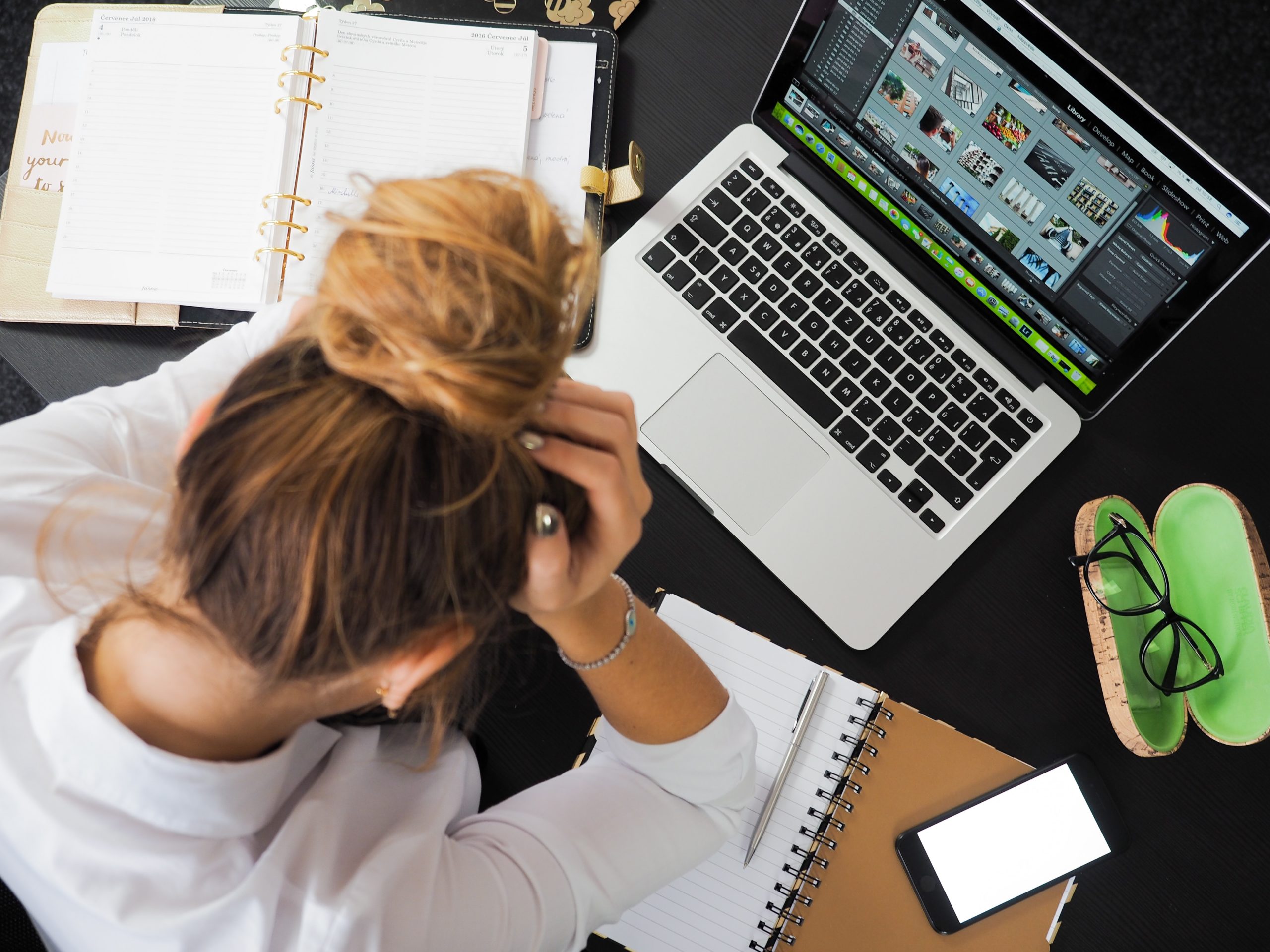 women with head down., hands on her head at her desk with anxiety. On her desk are an open planner, her laptop, glasses in their case , her cellphone and her notepad with a pen