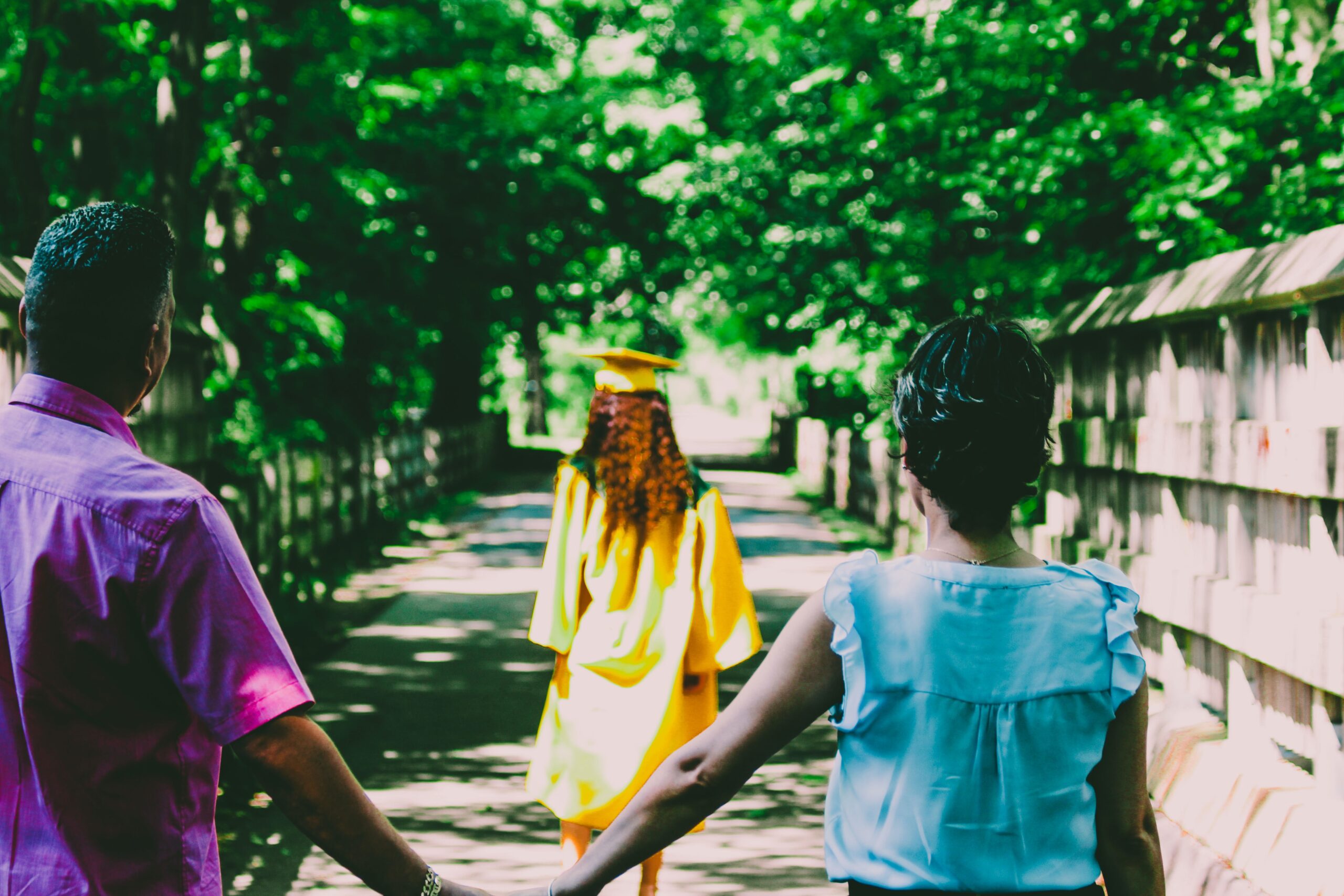 A woman in a yellow graduation cap and gown walking ahead of her parents who are holding hands